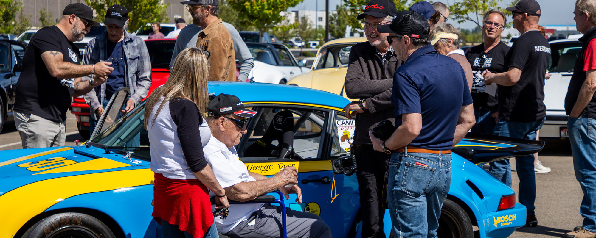 A crowd gathered around a blue and yellow classic Porsche 911 race car
